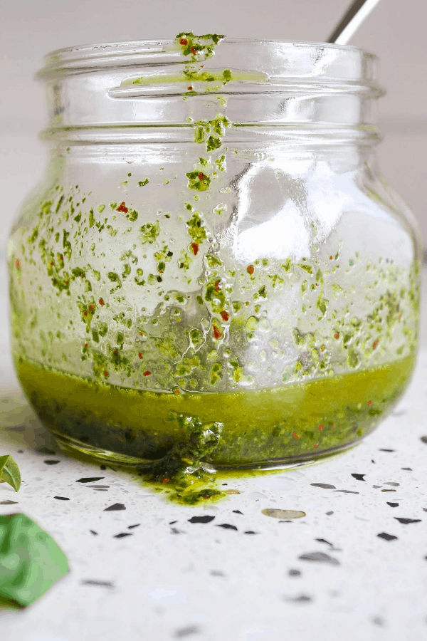Front shot of small jar of basil oil with spoon and sprig of basil on white surface. 