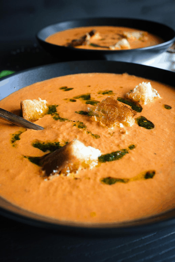 Front shot of two bowls of creamy tomato soup with grilled cheese croutons and basil oil. 