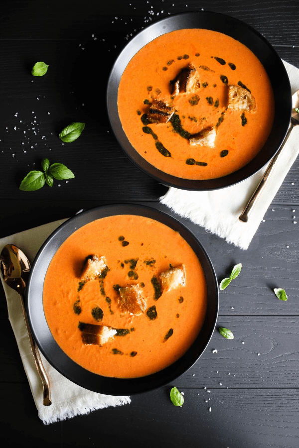 Top down shot of two bowls of Creamy Tomato Soup with Grilled Cheese Croutons & Basil Oil on grey surface with napkins, spoons, and scattered sea salt and basil leaves. 