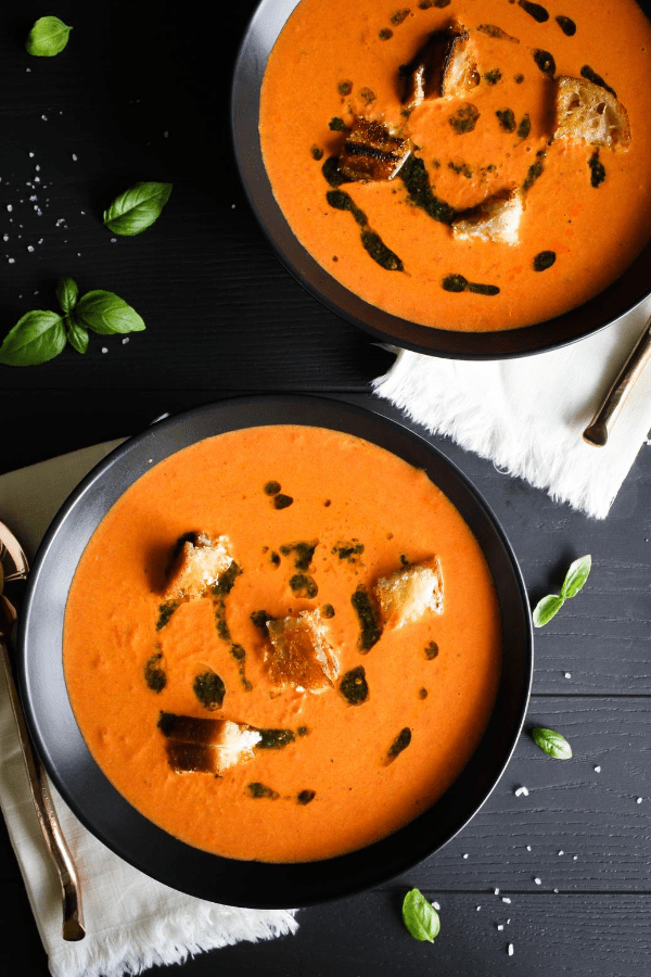 Top down shot of two bowls of creamy tomato soup with grilled cheese croutons and basil oil with napkins and spoons. 
