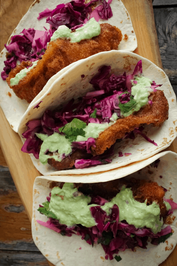 Close-up of three fish tacos w/ slaw and avocado salsa verdé on wood surface. 