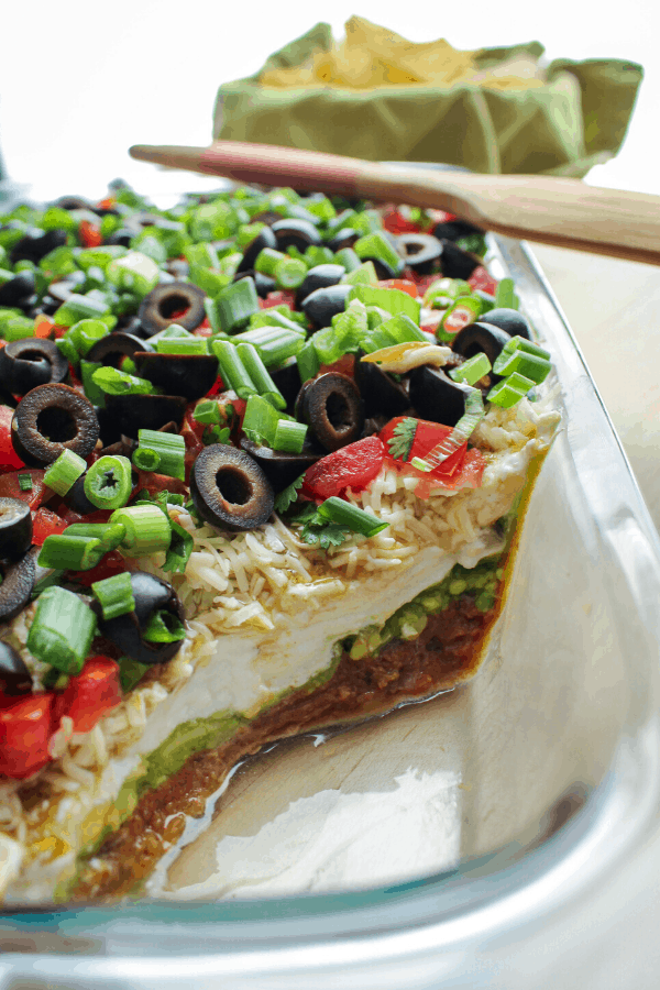 spicy 7 layer dip in a glass baking dish with wood spatula across and basket of chips in background