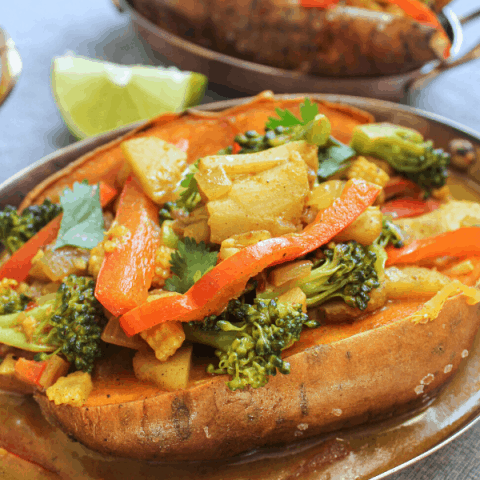 Two stuffed baked sweet potato topped with vegetable coconut curry in copper dish on blue surface with lime wedge