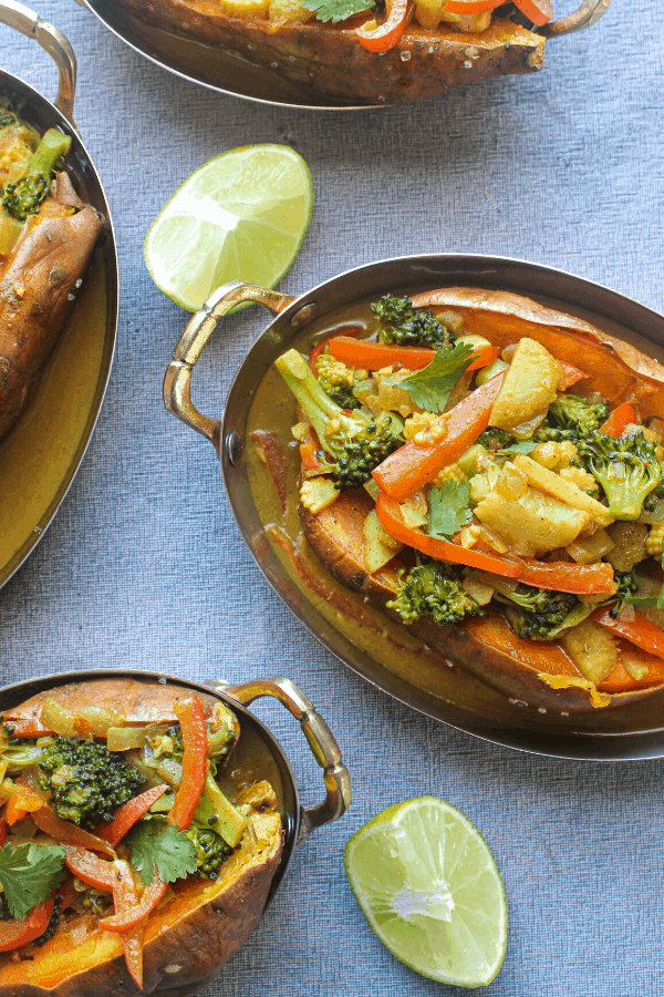 four stuffed sweet potatoes with vegetable coconut curry in copper dishes on blue surface with two lime wedges