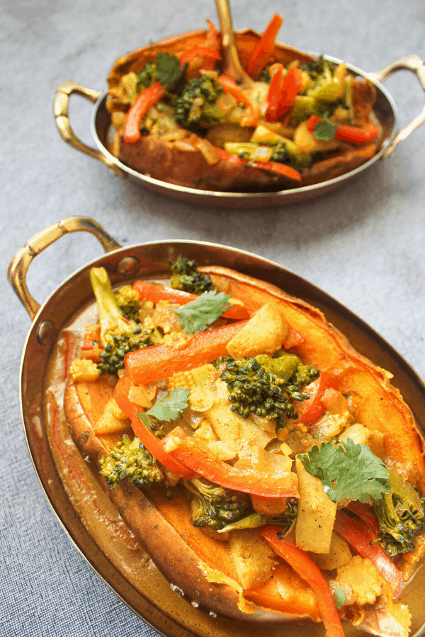 two stuffed sweet potatoes with vegetable coconut curry in copper dishes on blue surface 