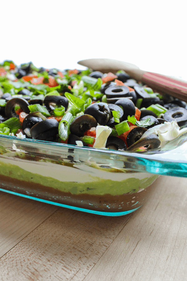 spicy seven layer dip in glass baking dish from the side with wooden spatula laying across corner