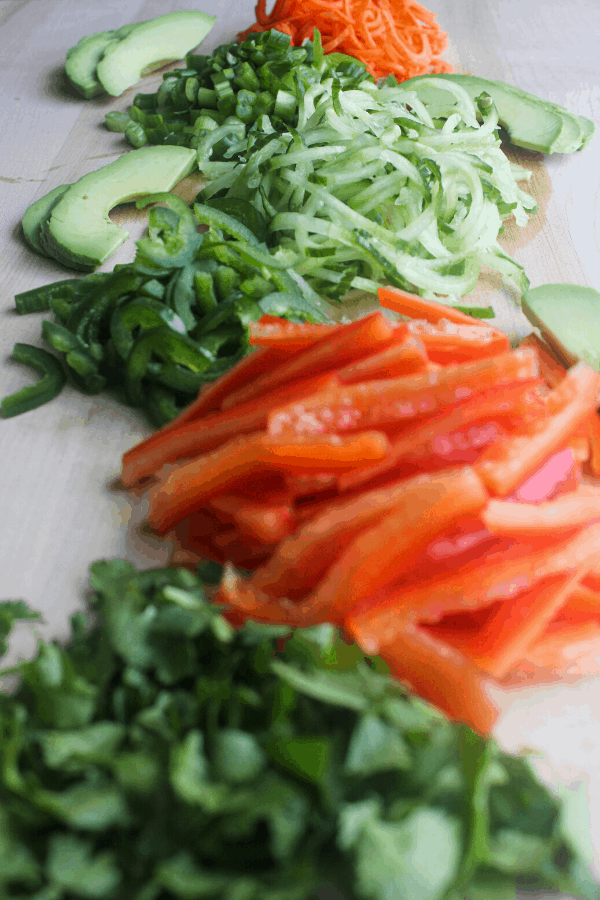 piles of carrots, green onions, cucumber, jalapenos, red bell pepper, and cilantro surrounded by sliced avocado on wood surface