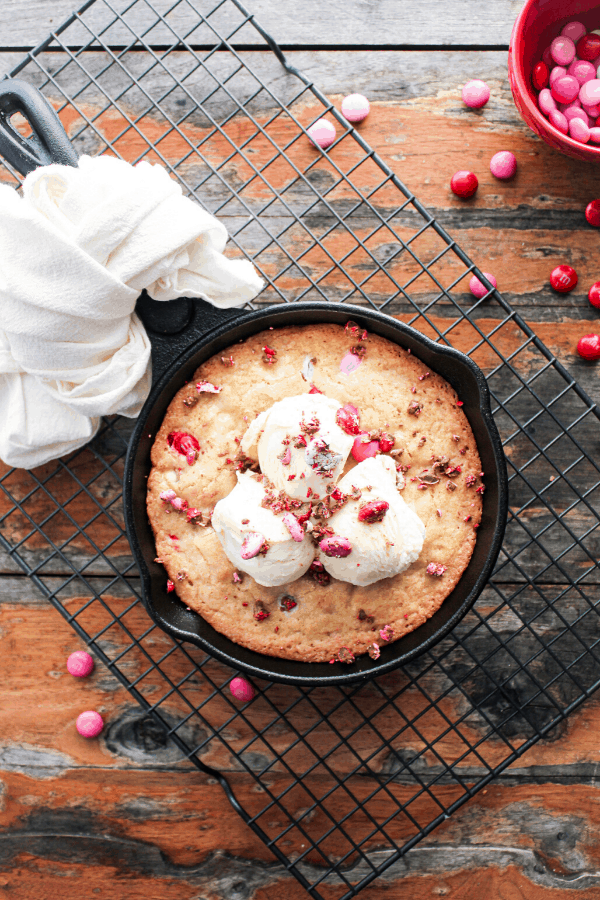 Valentine's blondie topped with three scoops of vanilla ice cream and crushed M&M's in acast iron skillet with white dish towel tied around handle on a wire cooling rack on a wood table with scattered M&M's and small red bowl of M&M's.