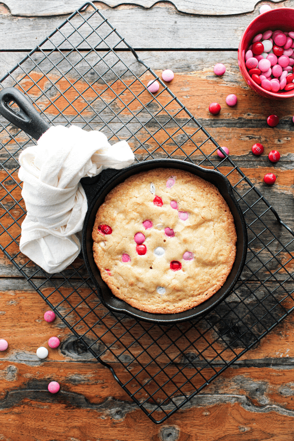 Valentine's blondie in a cast iron skillet with white dish towel tied around handle on a wire cooling rack on a wood table with scattered M&M's and small red bowl of M&M's.