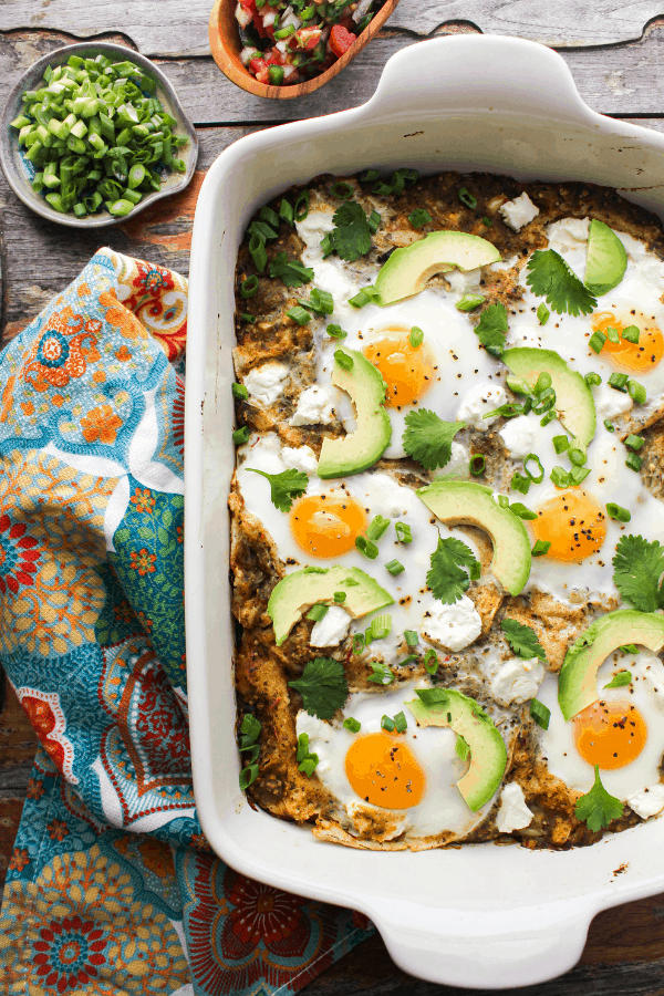 huevos rancheros casserole in white baking dish on wood table with blue patterned towel and small tray of green onion and bowl of pico de gallo from above. 