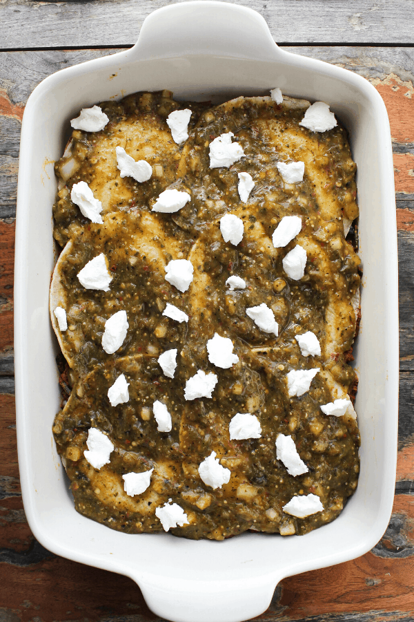 corn torillas covered in salsa verde and crumble goat cheese in white baking dish from above 