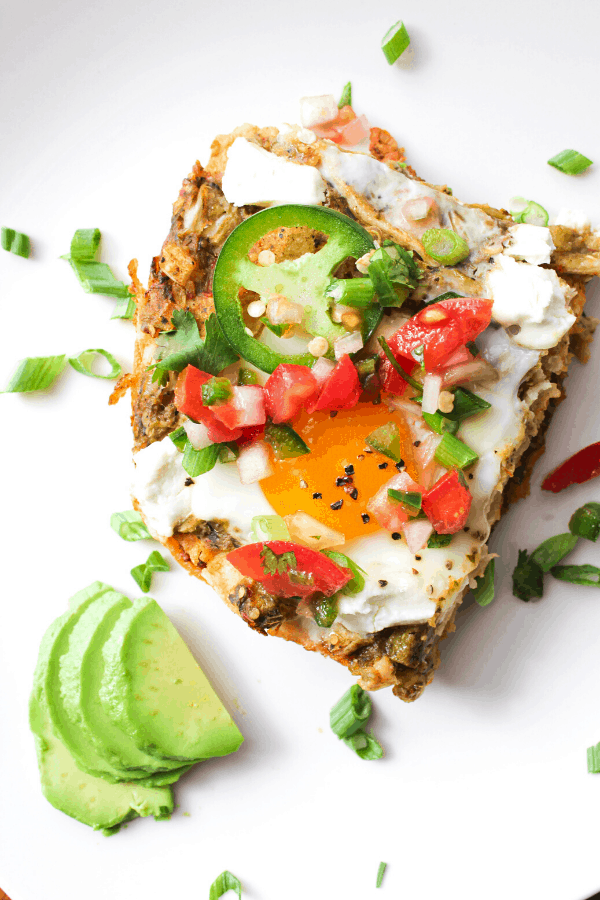 close up shot of slice of huevos rancheros casserole topped with pico de gallo and green onions on white surface with slices of avocado and 