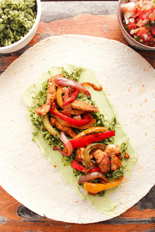 flat tortilla layered with chicken fajita wrap ingredients on table with small bowl of green rice and pico de gallo. 