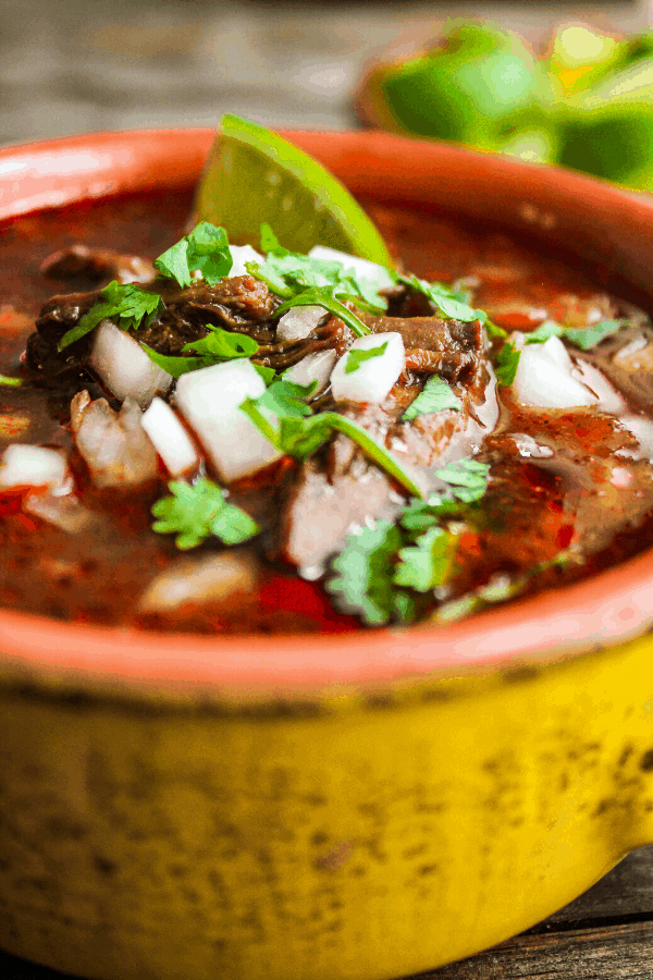 close up of beef birria in bowl topped with diced white onion, cilantro, and lime wedge with lime wedges in background 