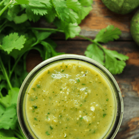 verde enchilada sauce in jar on table with bunch of cilantro and a tomatillo shot from above