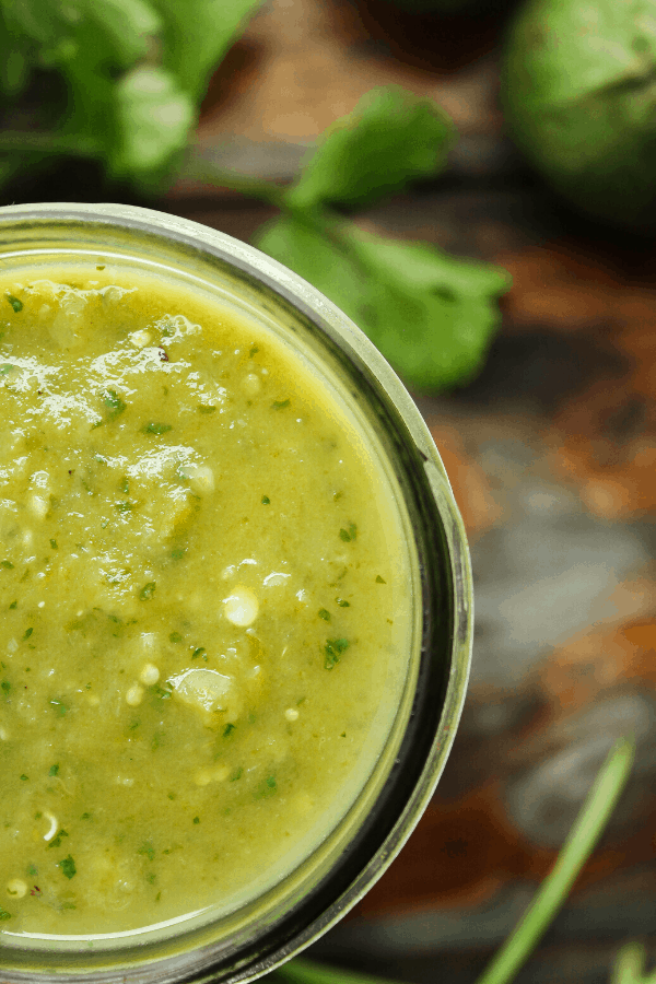 close up of verde enchilada sauce in jar with cilantro and tomatillo in background 