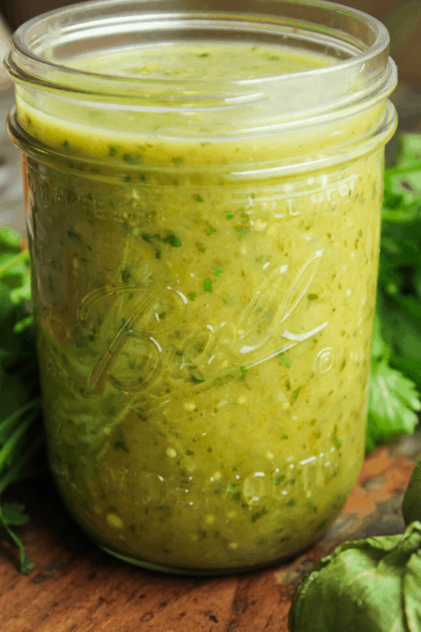 small jar of verde enchilada sauce with bunch of cilantro and a tomatillo on a distressed wood table 