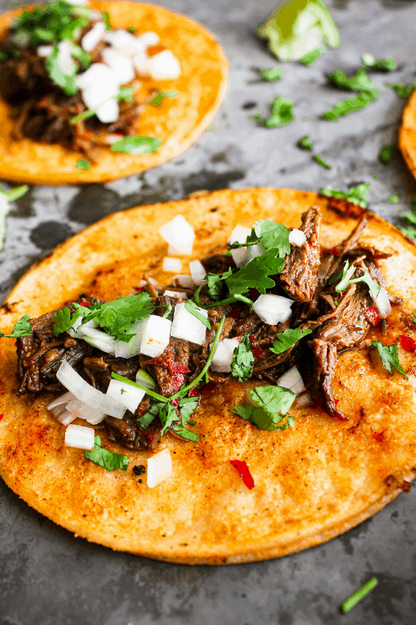 close up of beef birria taco with a second taco, lime wedge, and cilantro in the background