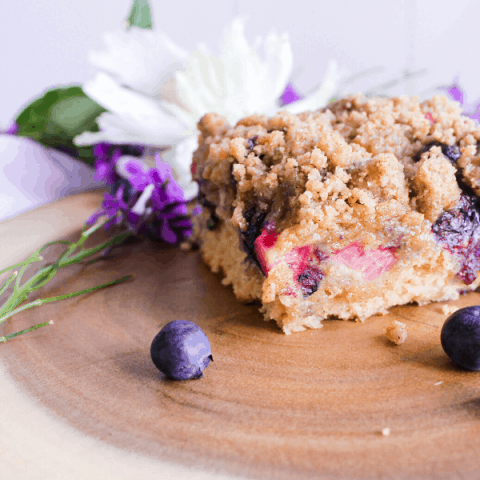 Slice of buckle cake on wood plater with scattered blueberries and flowers in background.
