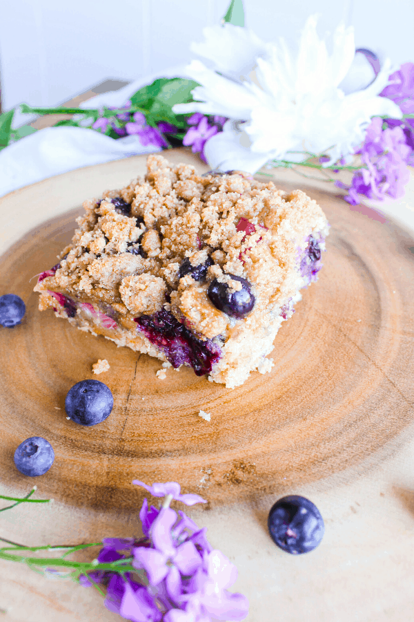 Slice of blueberry rhubarb buckle cake on wood trunk platter with scattered blueberries and flowers. 