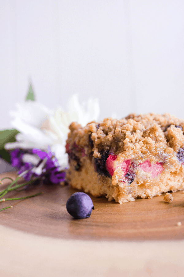 Close up side view of blueberry rhubarb buckle cake with a blueberry and flowers on a wood platter.