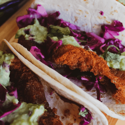 Close-up of three fish tacos w/ slaw and avocado salsa verdé on wood surface.