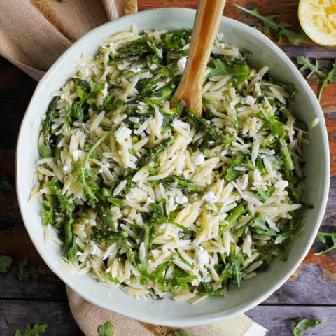 Large bowl of lemon orzo salad with wooden spoon atop a yellow towel with sprinkled arugula and lemon half shot from above.