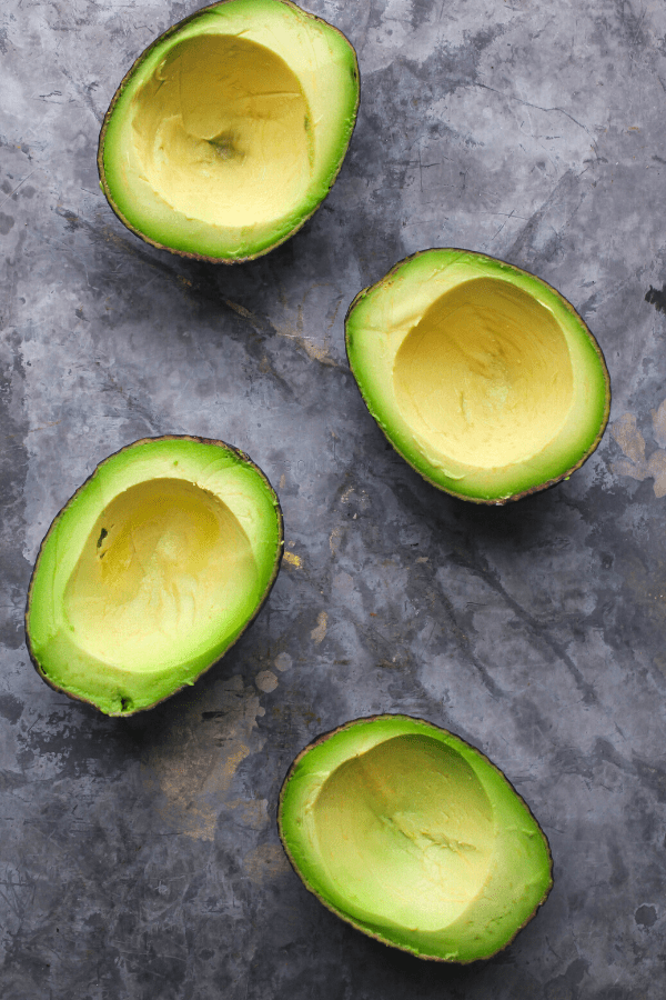 four pitted and scooped out avocado halves on a grey surface shot from above