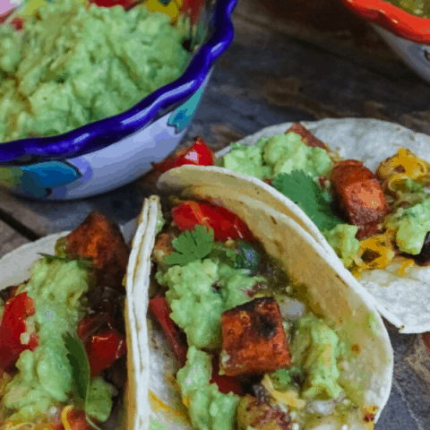 Three roasted vegetable tacos surrounding a small bowl of mashed avocado.