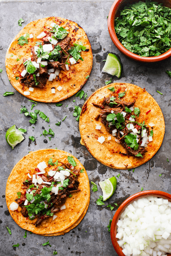three beef birria tacos laid flat on a sheet pan with sides of cilantro and white onions and scattered lime wedges shot from above