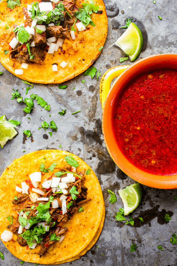 two beef birria tacos with bowl of consume on sheet pan with lime wedges and cilantro 