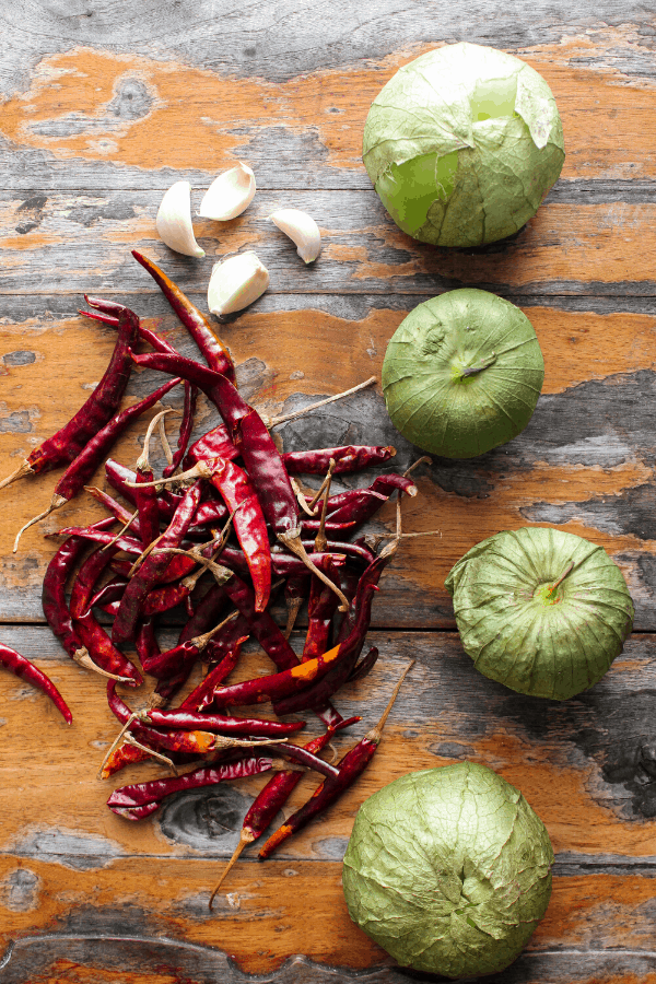 Four tomatillos, four garlic cloves, and pile of chile de árbol chiles on a distressed wood surface shot from above. 