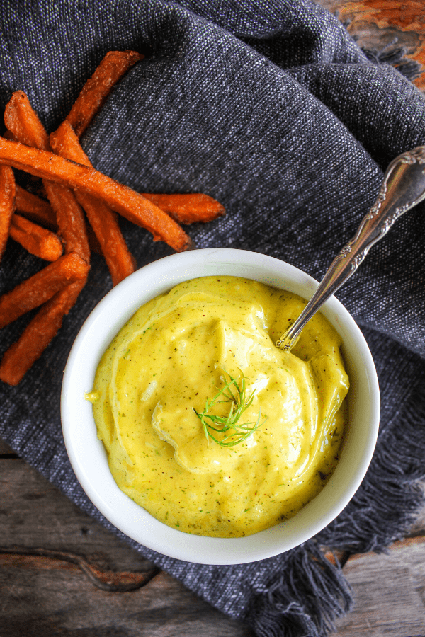 Small bowl of lemon dill aioli with a spoon atop a demin towel with pile of sweet potato fries. 