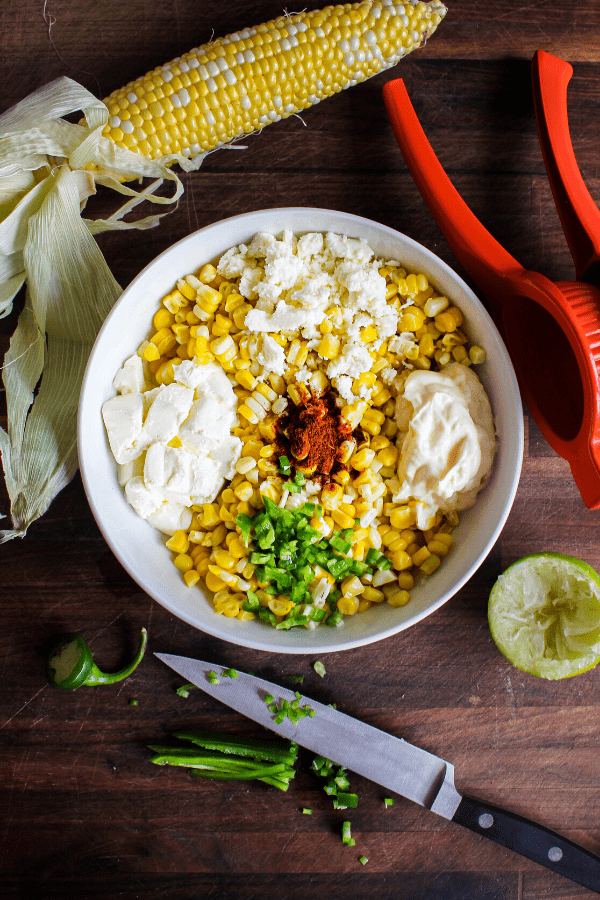 Mexican street corn ingredients in a white bowl with corn cob, juicer, lime, and diced jalapeno with knife on a wood surface shot from above. 