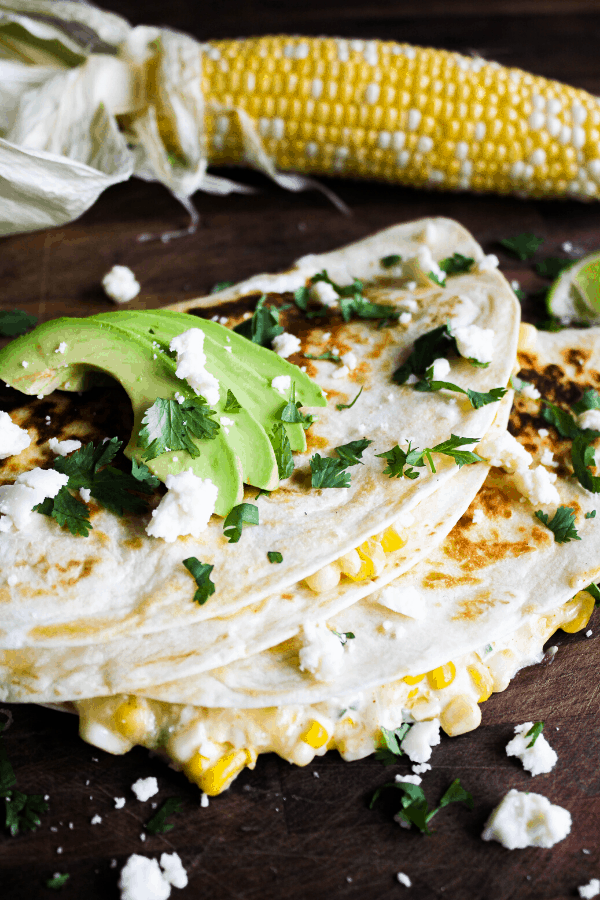 Two quesadillas stacked atop one another garnished with sliced avocado, cilantro, and Cotija cheese with a corn cob in the background. 