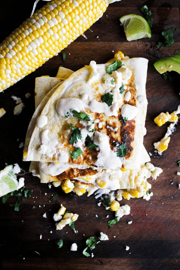 Slice Mexican street corn quesadillas stacked atop one another drizzled with creamy chipotle sauce and sprinkled with cilantro and Cotija cheese on table with corn cob and lime wedges.