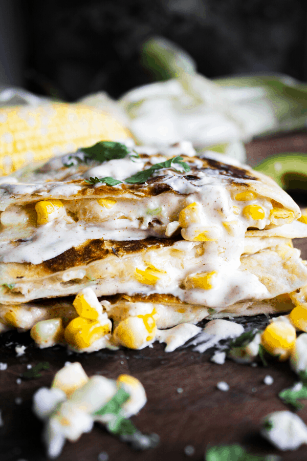 Close up of stacked quesadilla slices drizzled with creamy chipotle sauce with corn cob in background.