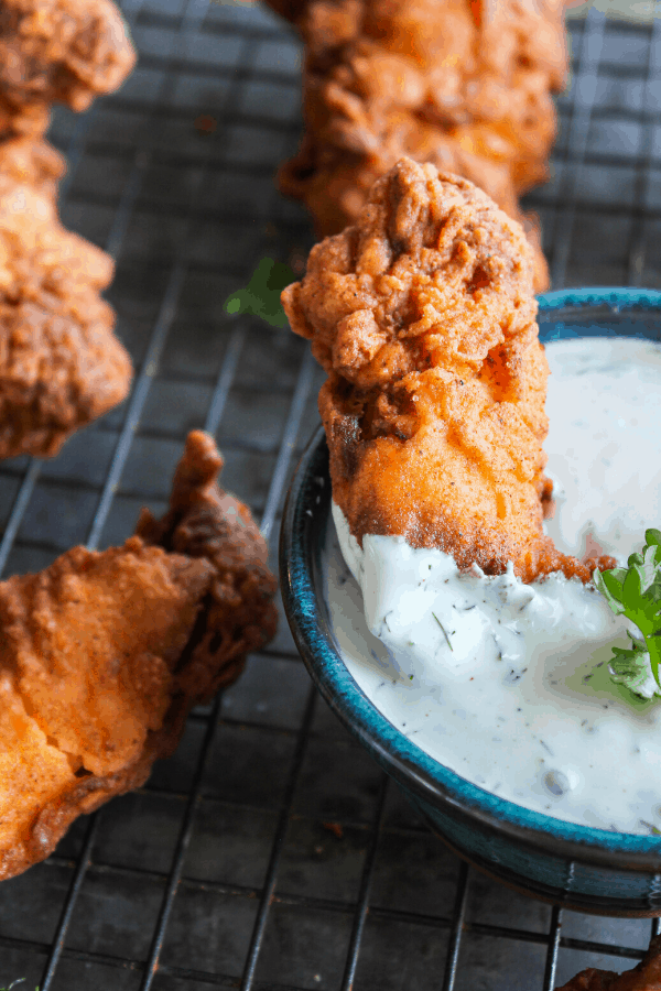 Fried chicken tender being dipped in blue bowl of dill ranch with additional chicken tenders in background on wire cooling rack. 