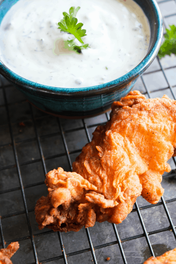 Close up of a fried chicken tender with dill ranch in small blue bowl on wire cooling rack. 