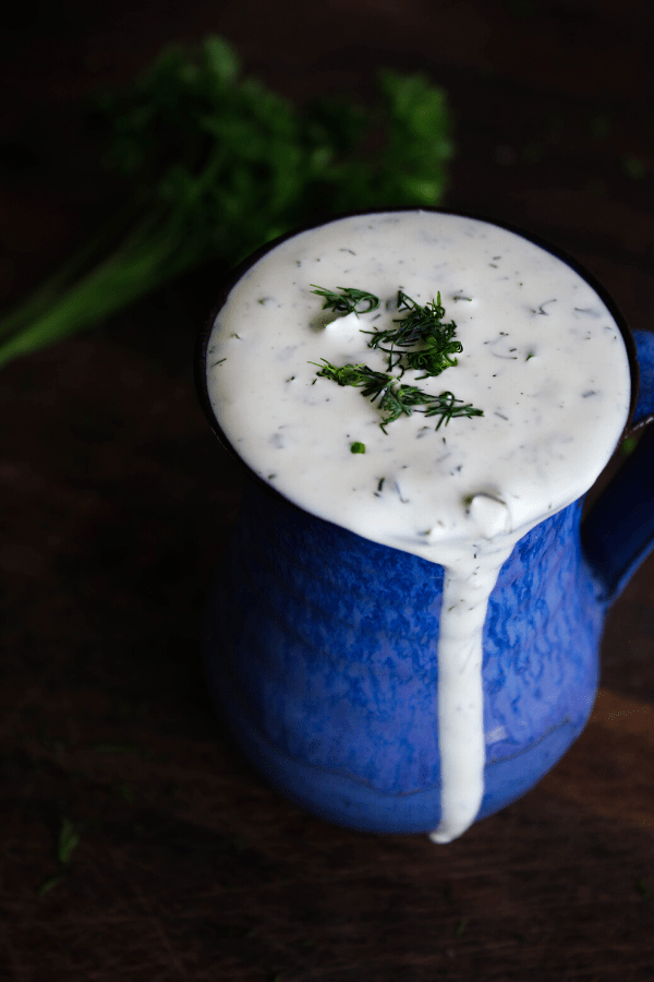 Ranch topped with fresh dill in blue vase atop dark wood surface with herbs in background. 