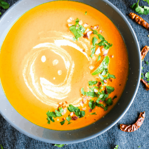 Bowl of Thai carrot soup in grey bowl on blue surface sprinkled with Thai chiles and cilantro.