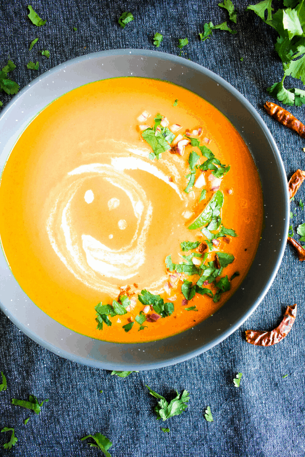 Bowl of Thai carrot soup in grey bowl on blue surface sprinkled with Thai chiles and cilantro.