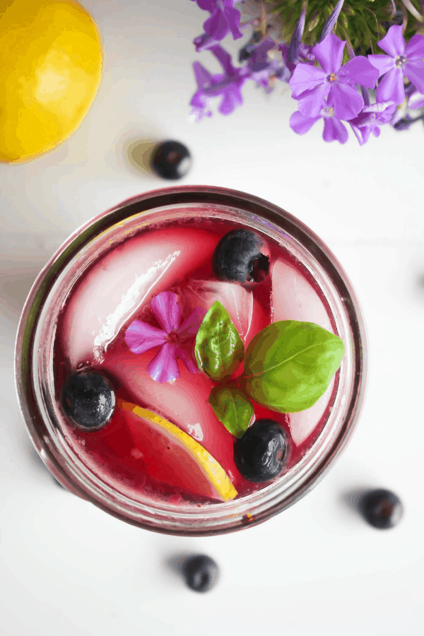Top down shot of jar of blueberry basil lemonade garnished with lemon slice, blueberries, basil, and a single purple flower on a white surface with scattered blueberries, a lemon, and purple flowers. 