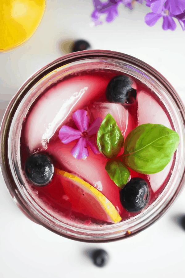 Close-up top down shot of blueberry basil lemonade garnished with blueberries, basil, lemon slice, and a single purple flower. 