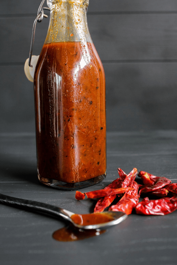 Bottle of chile de árbol hot sauce with spoon dripping with hot sauce and pile of chile peppers. 