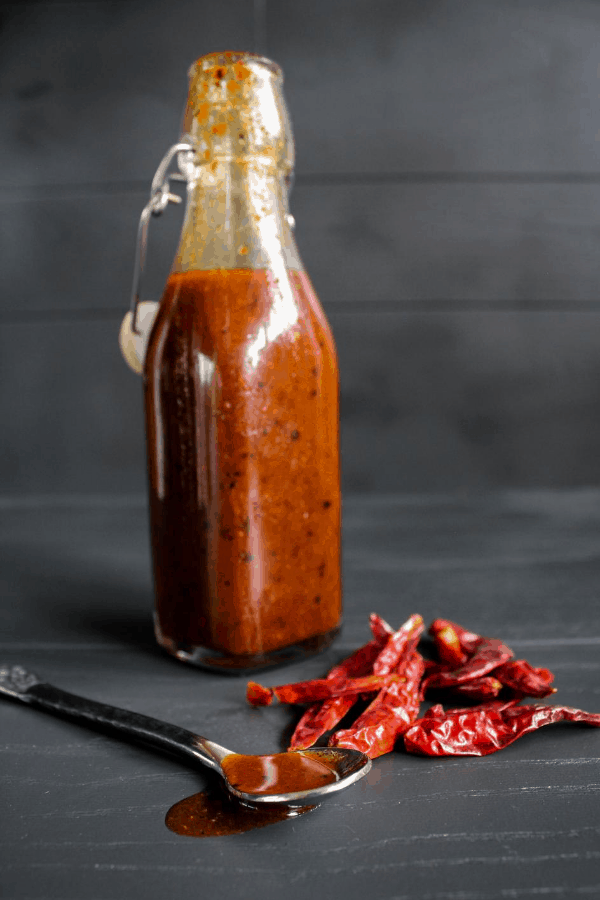 Bottle of hot sauce with a pile of chile peppers and a spoon dripping with hot sauce. 