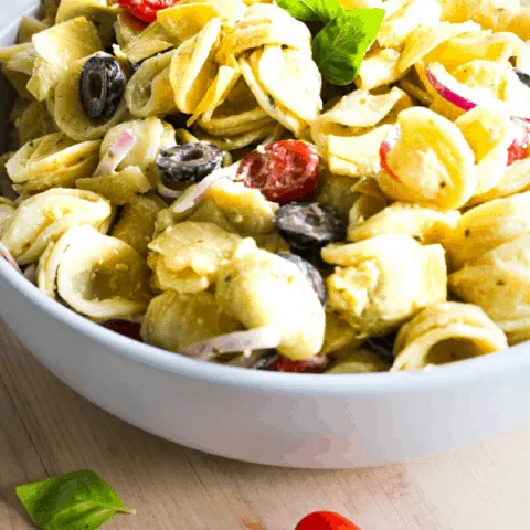Front shot of bowl of creamy pesto pasta salad on wood surface with three grape tomatoes and a basil leaf.