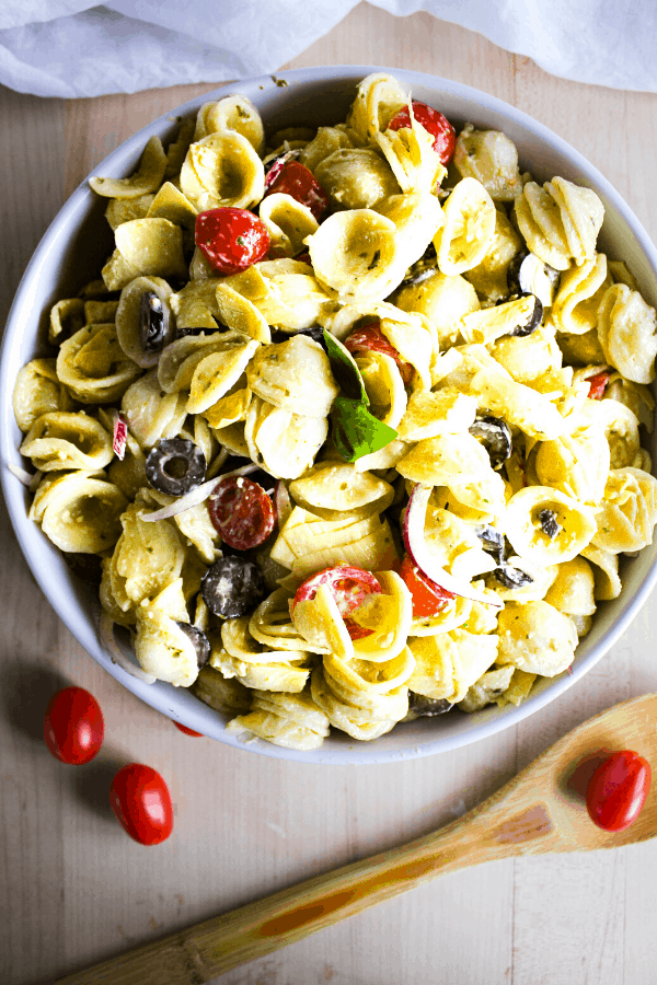 Large white bowl of creamy pesto pasta salad shot from above with wooden spoon and grape tomatoes. 