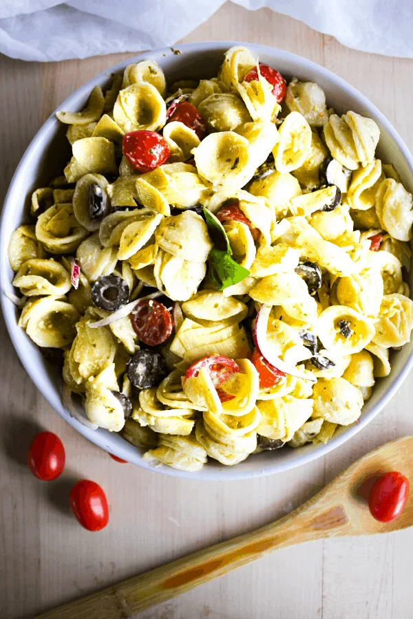 Large white bowl of creamy pesto pasta salad shot from above with wooden spoon and grape tomatoes.