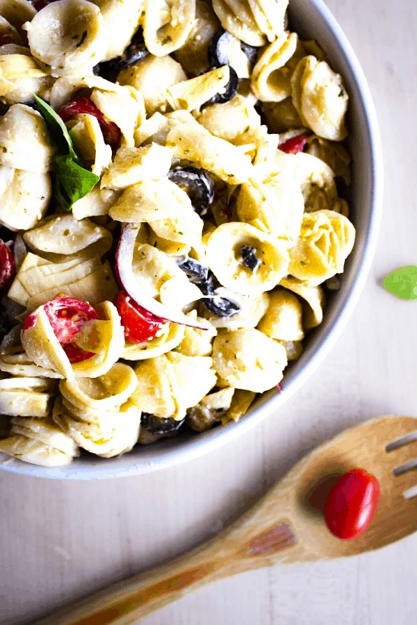 White bowl of pesto pasta salad on wood surface with a wooden spoon topped with a grape tomato shot from above.